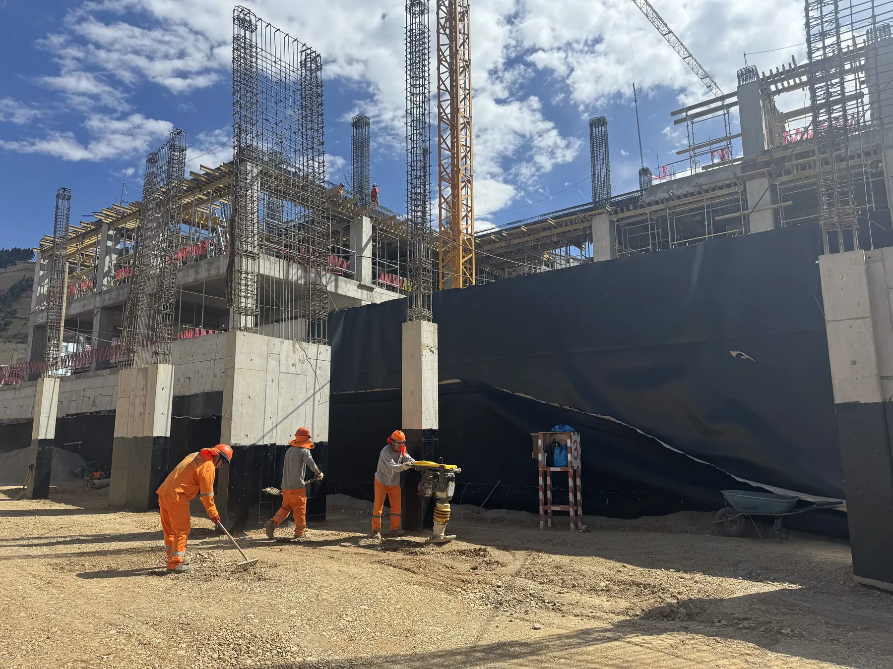 Obra de un hospital en construcción: armaduras de columnas montadas sobre muros de concreto, encofrados en los pisos superiores, una grúa torre al fondo y tres trabajadores nivelando el terreno.